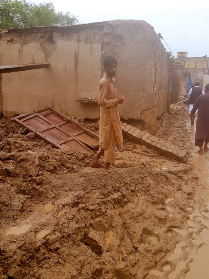 A street left under mud after floodwater receded, with a collapsed boundary wall behind.